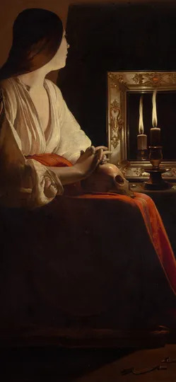 A woman sits in near darkness beside a candle and its reflection in a mirror, her hands resting on a skull in her lap.